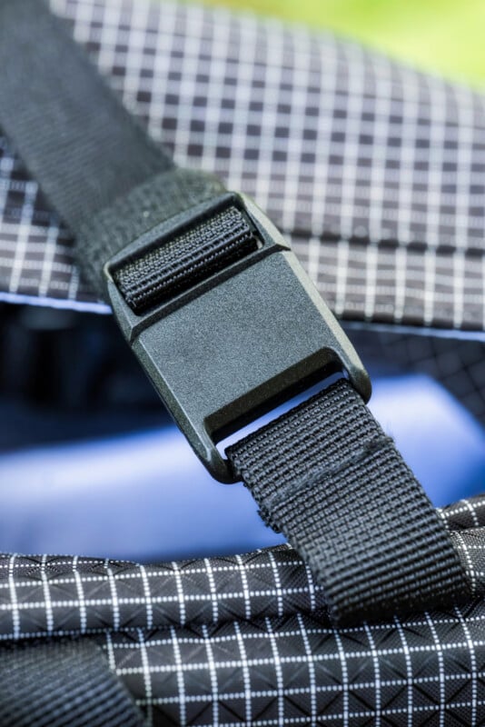 A close-up of a black plastic buckle fastened on a strap, attached to fabric with a black and white grid pattern, likely part of a backpack or bag.