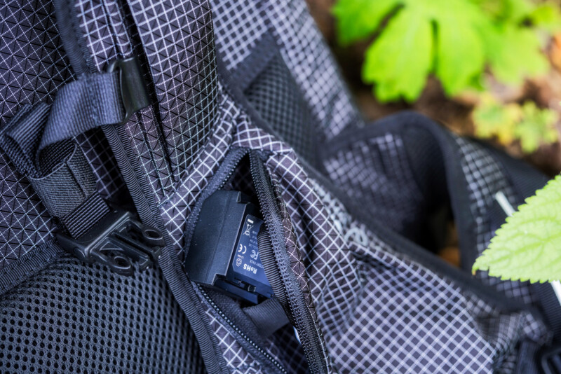 A close-up of a black and gray mesh backpack pocket partially unzipped, revealing a camera battery inside. Green leaves are visible in the background.