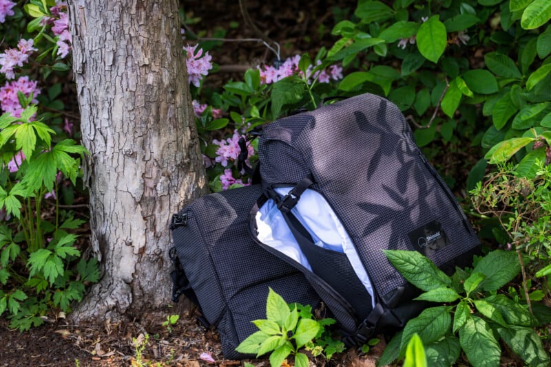 A black and gray backpack and a white shirt rest against a tree trunk, surrounded by green leaves and pink flowers in a garden setting.