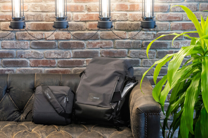 Two black bags rest on a dark leather couch against a brick wall with three industrial-style lights above. A green plant is on the right side of the couch.