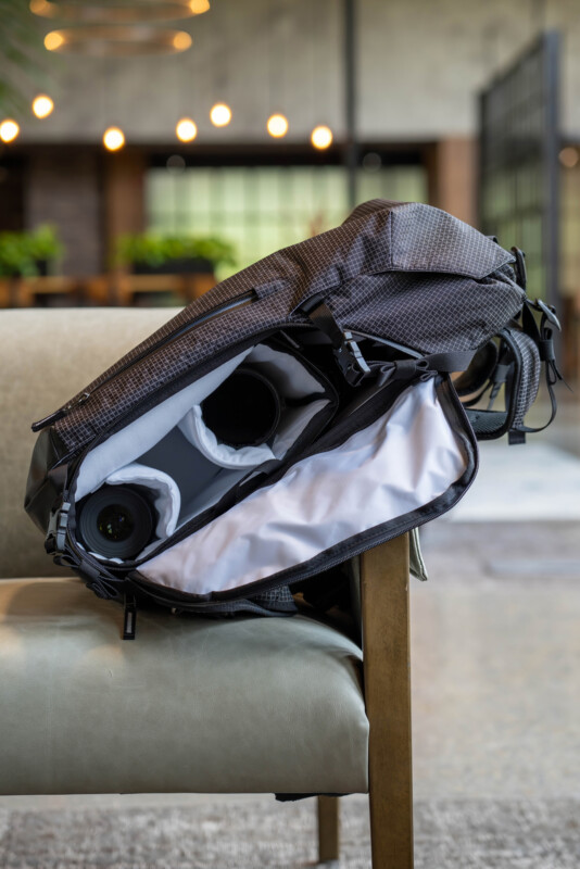 A gray camera backpack sits open on a chair, revealing padded compartments holding camera gear, including a camera body and a lens, in a modern indoor setting with blurred lights and greenery in the background.