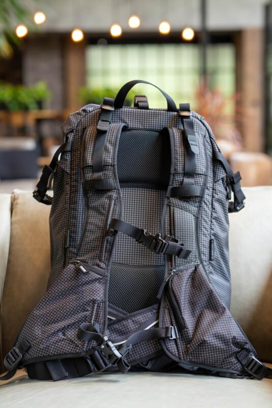 A gray and black hiking backpack with padded straps and multiple buckles sits on a light-colored couch indoors, with a blurred background of plants and hanging lights.