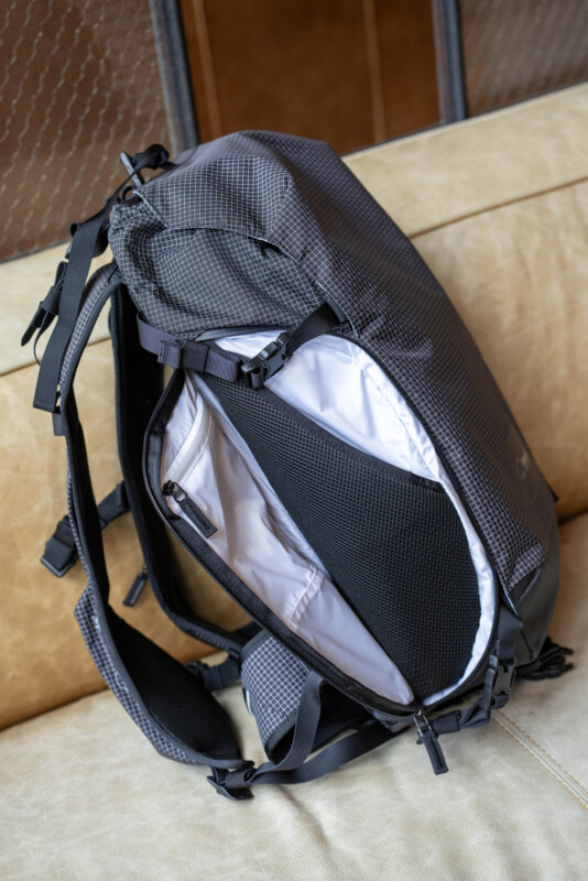 A black backpack with a white interior lining sits open on a beige couch, showing mesh pockets and organized compartments inside.