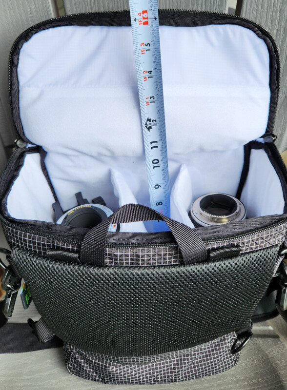 An open camera bag with a white interior, two camera lenses inside, and a tape measure standing vertically, showing the bag's depth. The exterior of the bag is dark with grid patterns.