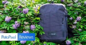 A black, grid-patterned Shimoda backpack sits among lush green leaves and purple flowers. Overlay text reads "PetaPixel Reviews" in the lower left corner.