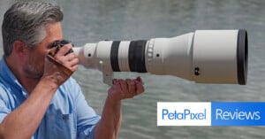 A man in a blue shirt takes a photo with a large telephoto camera lens near water. The image features the Petapixel Reviews logo in the bottom right corner.