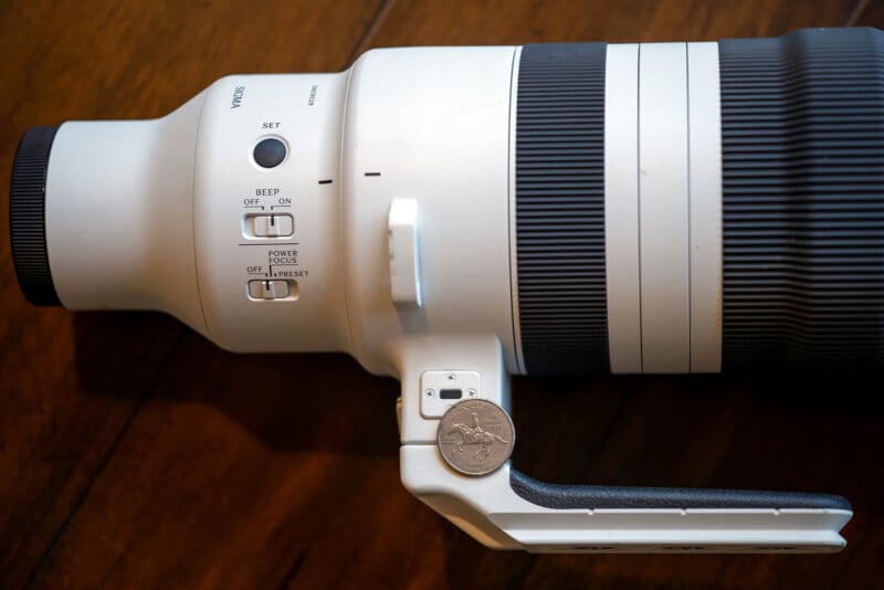 A large white telephoto camera lens with several switches and a mounting bracket is shown resting on a wooden surface. A U.S. quarter is placed near the mount for scale.