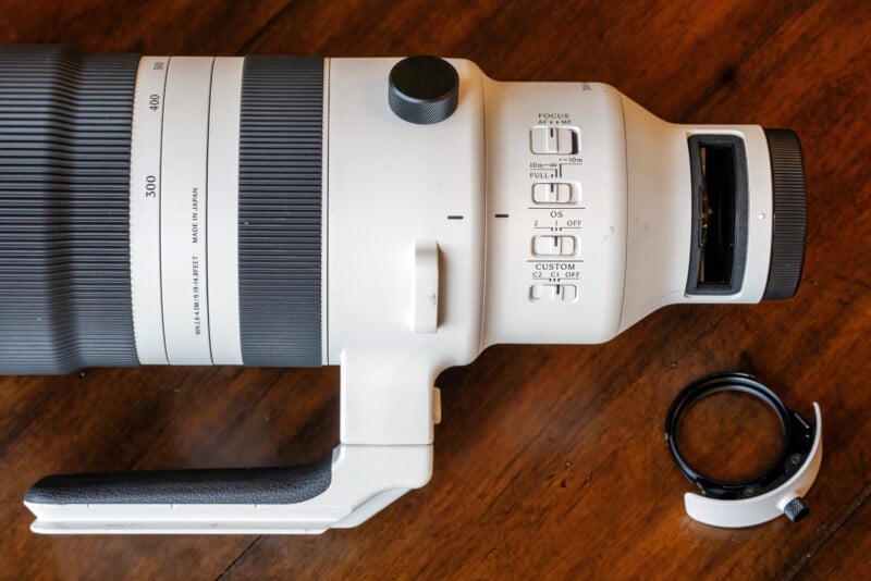 A large white telephoto camera lens with a handle lies on a wooden surface, showing focus and stabilization switches. A detached lens collar ring is placed nearby.