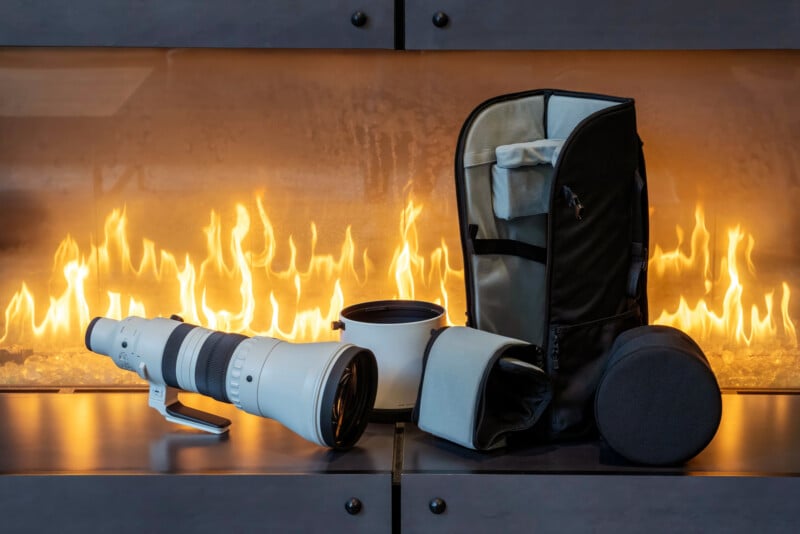 A large white telephoto camera lens, a lens hood, a round lens case, and a black carrying bag are displayed on a surface in front of a modern fireplace with bright flames.