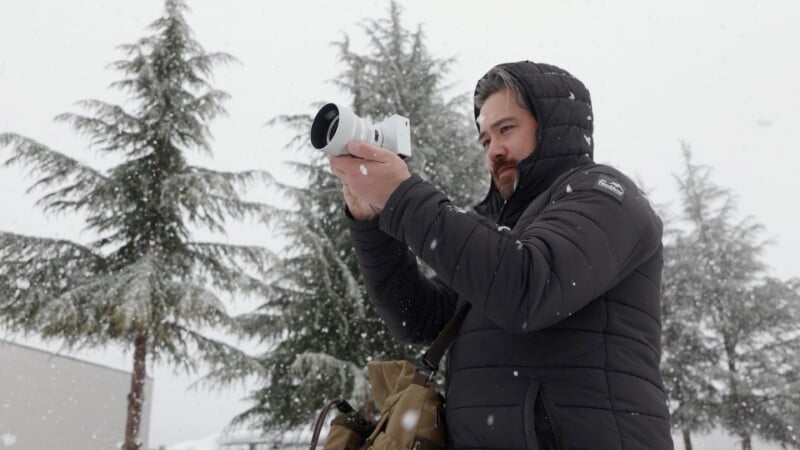 A person in a black winter jacket holds a camera while standing outdoors in snowy conditions. Tall evergreen trees in the background are dusted with snowflakes. The person appears focused on capturing a photo.