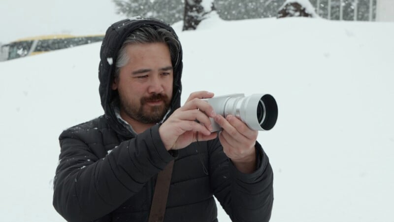 A man in a black hooded jacket is taking photos in a snowy landscape. He is looking at the camera in his hands, surrounded by snow-covered trees.