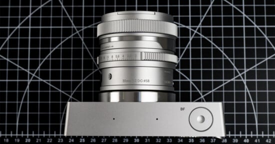 A modern silver camera lens with detailed markings, positioned vertically on a silver surface. The background features a black and white grid pattern with circular and diagonal lines.