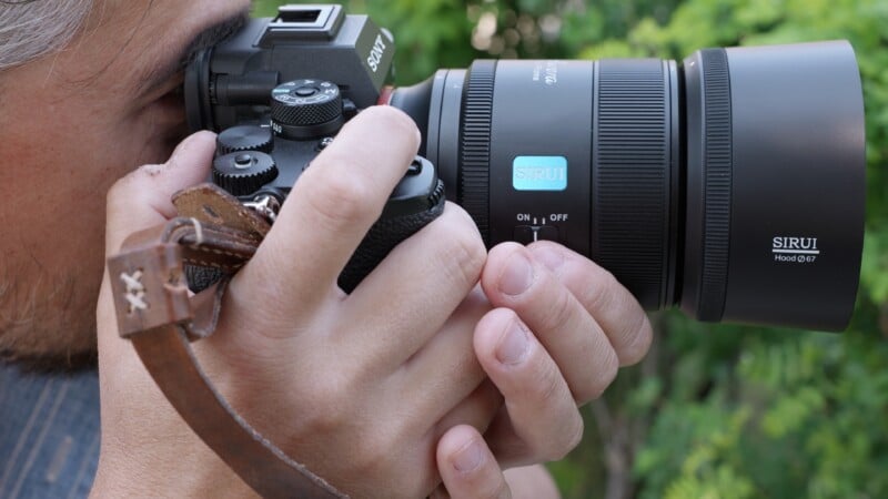 A close-up of a person holding a black digital camera with a Sirui lens, focusing on the camera and hands against a blurred green outdoor background.