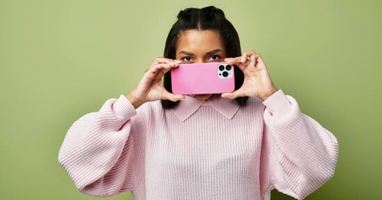 A person in a light pink sweater holds a pink smartphone horizontally, partially covering their face, against a plain light green background.