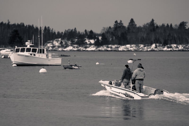 Two people in a small motorboat head towards a larger boat named "Hamley House" on a calm body of water. Surrounding trees are visible in the distance, and the scene is in black and white.