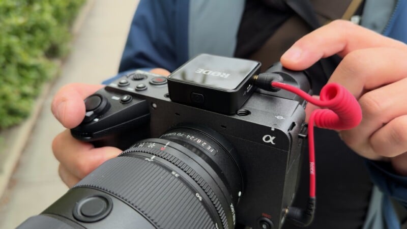 A person’s hands holding a Sony Alpha camera with a Rode wireless microphone attached, adjusting the top-mounted device; the background shows greenery and a sidewalk.
