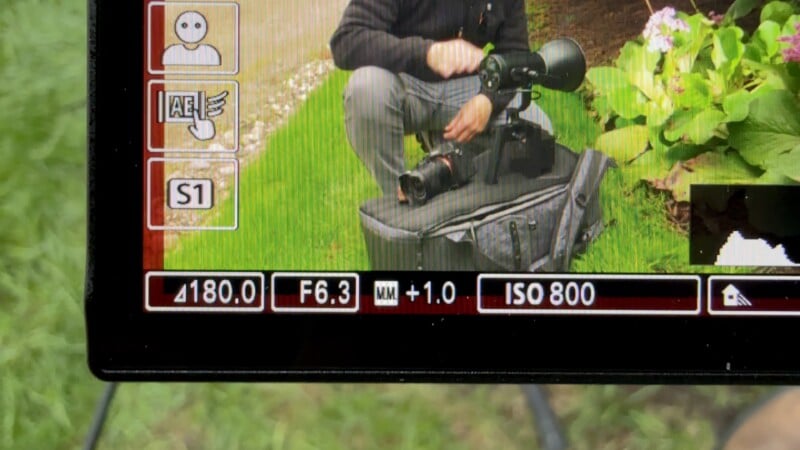 A camera screen displays settings like 4180.0, F6.3, +1.0, and ISO 800, with a man kneeling outside holding a telephoto lens and camera, partially visible behind camera interface icons.