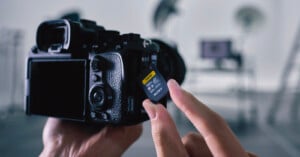 A person inserts a Sony SD memory card into a digital camera in a studio setting, with blurred lights and equipment in the background.