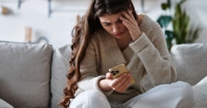 A woman with long brown hair sits on a couch, looking stressed as she holds a smartphone in one hand and rests her other hand on her forehead. She appears worried or upset.