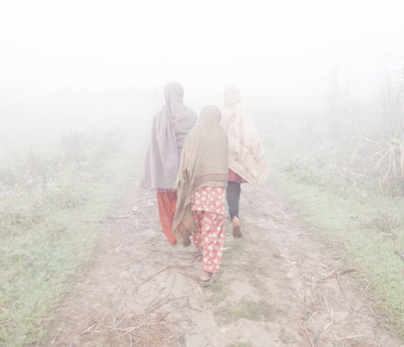 Three women in traditional clothing walk down a dirt path surrounded by grass, shrouded in dense white fog. The mist obscures the background, creating a soft, ethereal atmosphere.