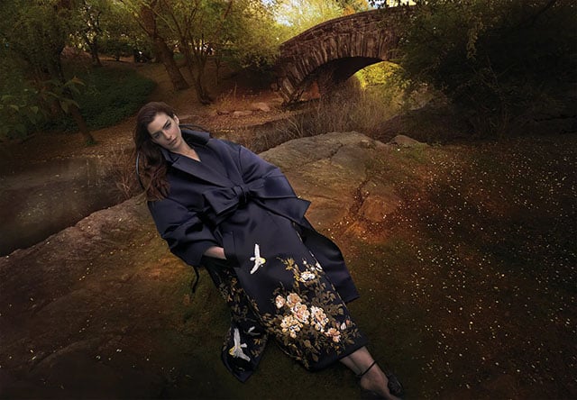 A person in a dark, elegant robe with floral and bird designs sits on a rock in a serene, wooded area near an old stone bridge, surrounded by soft, golden light and greenery.