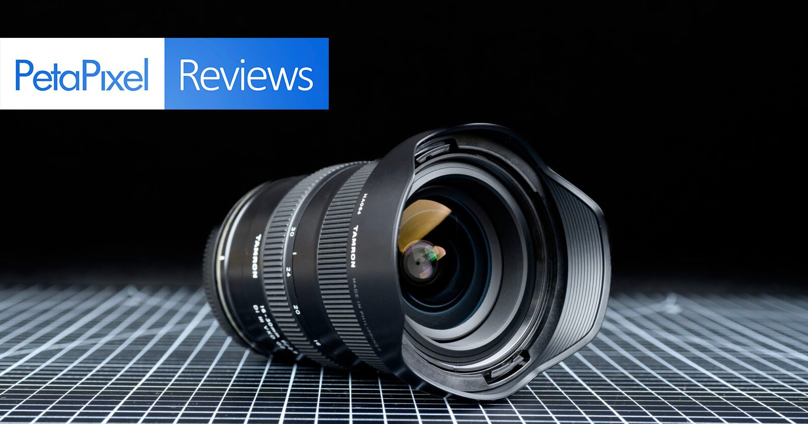 A wide-angle camera lens lies on a grid-patterned surface with a black background. The top left corner displays the "PetaPixel Reviews" logo in blue and white.