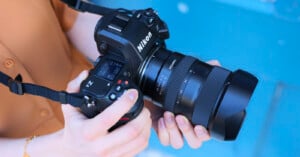 A person holds a Nikon Z series camera with a Sigma lens attached, against a blurred blue background. The camera is positioned for shooting, and the person's hands are visible adjusting the controls.