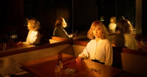 A woman with blond hair sits alone at a dimly lit booth, her reflection appearing multiple times in mirrors behind her. The table has a glass, an ashtray, and salt and pepper shakers. The mood is contemplative and moody.