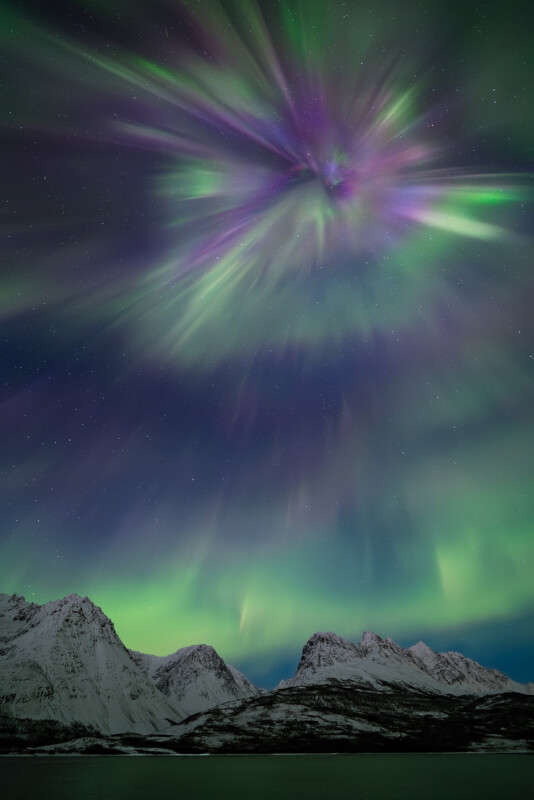 Aurora borealis in green and purple hues lights up the night sky above snow-covered mountains and a calm body of water.