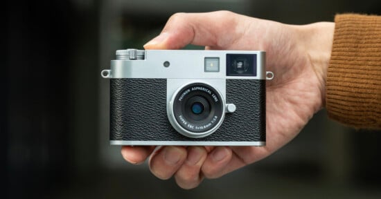 A hand holding a compact, retro-style digital camera with a silver and black body, textured grip, and lens facing forward against a blurred background.