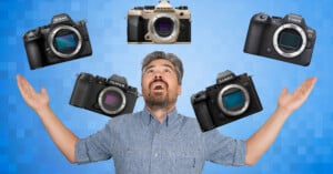 A man with a beard and gray hair stands with arms raised, looking upward, as five mirrorless cameras from different brands float above him against a blue, checkered background.