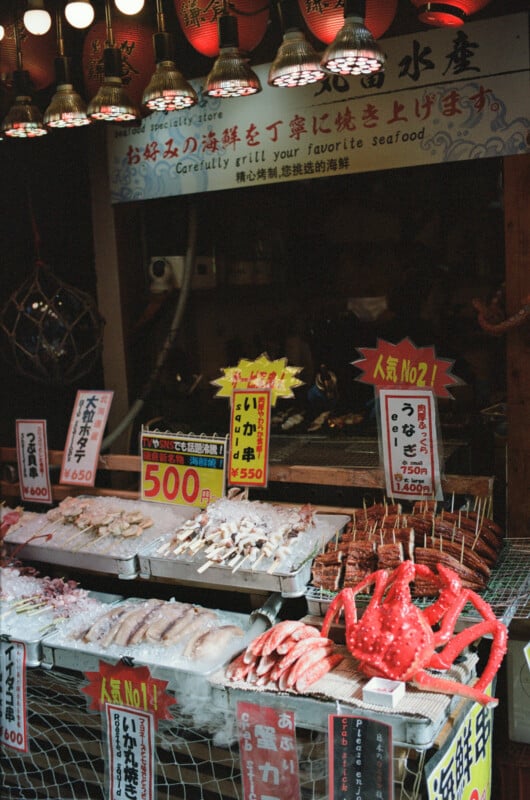A seafood market stall displays various fresh seafood, including skewered squid, shrimp, and a large red crab, with colorful signs and Japanese text above and on the stall. Red lanterns hang overhead.