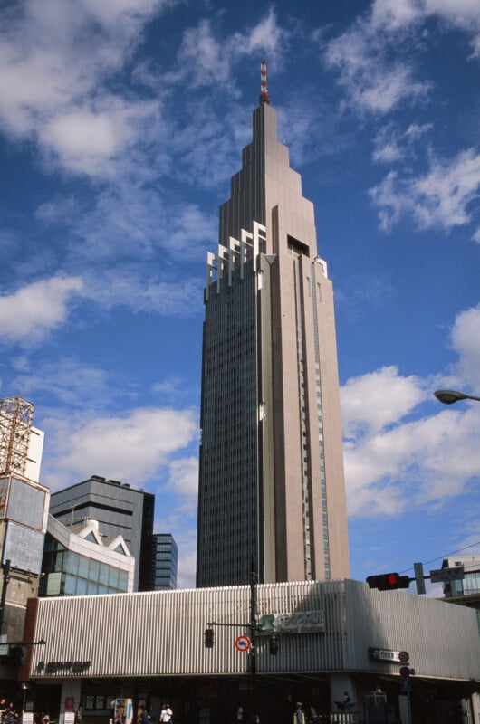 A tall, modern skyscraper with a tiered, stepped design rises against a blue sky with scattered clouds, surrounded by shorter buildings at its base in an urban setting.