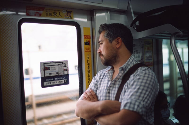 A man with gray hair and a beard, wearing a plaid shirt and shoulder bag, stands with arms crossed, looking out the window of a train with Japanese signs and stickers on the walls.