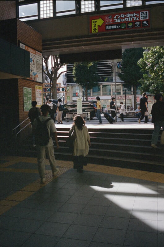 People walk up steps toward a sunlit outdoor area from a shaded indoor space. Trees, buildings, and signs in Japanese are visible outside. Some people carry bags, and shadows are cast on the tiled floor.