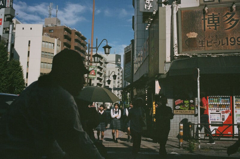 Silhouetted person in foreground with students in uniforms walking on a city street in Japan; buildings, signs, and a vending machine are visible under a sunny blue sky.