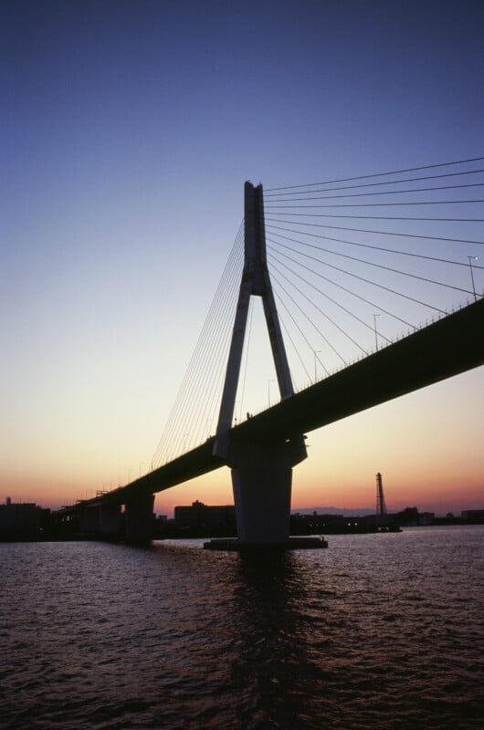 A cable-stayed bridge spans a body of water at sunset, with the sky fading from orange to deep blue and city silhouettes visible in the background.