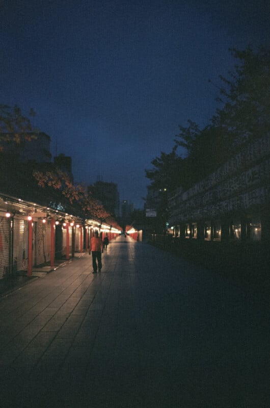 A person walks alone down a dimly lit, empty street at night, with small lights lining the storefronts on either side and trees silhouetted against the dark blue sky.