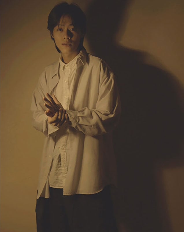A person stands indoors against a beige wall, wearing a loose white shirt over a buttoned top. Soft, warm lighting casts a large shadow of the person on the wall behind them.