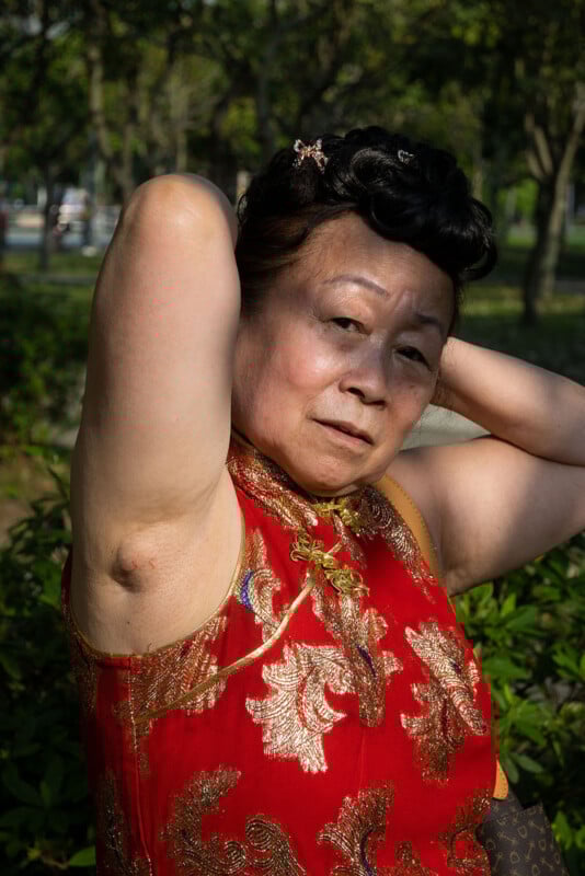 An older woman in a red, gold-patterned traditional dress poses outdoors, arms raised behind her head. She has styled black hair and a serious expression. Green foliage and trees are visible in the background.