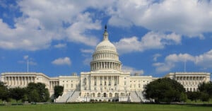 The United States Capitol building with its white dome and columns, set against a blue sky with scattered clouds and surrounded by green lawns and trees.