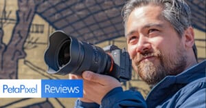 A person with a beard smiles and holds a camera with a large lens, standing outdoors in front of a mural. The image includes the text "PetaPixel Reviews" in the lower left corner.