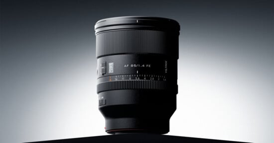 A black camera lens with a focus ring and aperture markings stands upright on a dark surface against a gradient gray background, illuminated from behind.