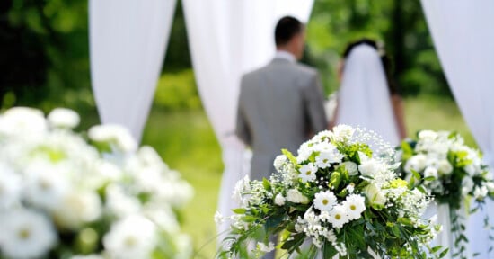 A bride and groom stand together outdoors under a white arch, with their backs to the camera. White floral arrangements and draped fabric decorate the scene, suggesting a wedding ceremony.
