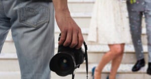 A person in jeans holds a camera, standing on steps. In the blurred background, two people dressed in formal attire, possibly a bride and groom, are descending the stairs.