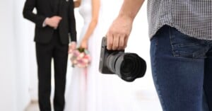 A person in jeans holding a camera stands in the foreground, while a bride in a white dress and a groom in a black suit stand together, slightly out of focus, in the background.